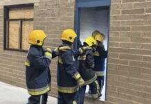 4 policing students in turnout overalls enter a derelict training building lit by torchlight