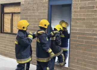 4 policing students in turnout overalls enter a derelict training building lit by torchlight