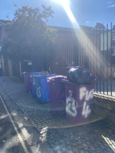 Row of 5 overflowing purple and blue bins outside a building in Liverpool. Photo copyright - Hannah O'Sullivan (BA Journalism 2025)