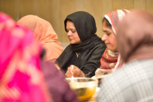 Image shows group of women sitting together, engaged in a communal meal. They are wearing colourful headscarves and traditional clothing. 
