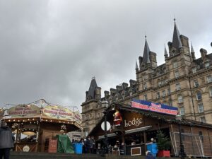 The photo shows an outdoor Christmas market set against a large, ornate stone building with many pointed towers and decorative spires. The sky above is grey and overcast, giving everything a wintry feel.In the foreground, there are two wooden market stalls. On the left is a round stall with a sign that reads “Swing Grill German Sausage.” Warm yellow lights are strung around its roof, giving it a cozy glow. A few people are standing nearby, bundled up in coats. To the right is a larger wooden cabin-style stall with a sign that says “Lodge.” Above its roof hangs a big banner reading “Welcome to Liverpool Christmas Markets” in red and blue lettering. A life-size Santa figure is mounted just under the sign. There are small Christmas decorations along the roofline, and a tiny Christmas tree sits outside the stall. A handful of people are walking around the market area, wearing winter jackets. Some plastic bins and coverings are visible around the stalls, suggesting setup or storage. The scene feels festive despite the grey weather, with warm lights contrasting the cloudy sky. 