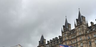 The photo shows an outdoor Christmas market set against a large, ornate stone building with many pointed towers and decorative spires. The sky above is grey and overcast, giving everything a wintry feel. In the foreground, there are two wooden market stalls. On the left is a round stall with a sign that reads “Swing Grill German Sausage.” Warm yellow lights are strung around its roof, giving it a cozy glow. A few people are standing nearby, bundled up in coats. To the right is a larger wooden cabin-style stall with a sign that says “Lodge.” Above its roof hangs a big banner reading “Welcome to Liverpool Christmas Markets” in red and blue lettering. A life-size Santa figure is mounted just under the sign. There are small Christmas decorations along the roofline, and a tiny Christmas tree sits outside the stall. A handful of people are walking around the market area, wearing winter jackets. Some plastic bins and coverings are visible around the stalls, suggesting setup or storage. The scene feels festive despite the grey weather, with warm lights contrasting the cloudy sky.