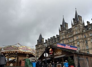 The photo shows an outdoor Christmas market set against a large, ornate stone building with many pointed towers and decorative spires. The sky above is grey and overcast, giving everything a wintry feel. In the foreground, there are two wooden market stalls. On the left is a round stall with a sign that reads “Swing Grill German Sausage.” Warm yellow lights are strung around its roof, giving it a cozy glow. A few people are standing nearby, bundled up in coats. To the right is a larger wooden cabin-style stall with a sign that says “Lodge.” Above its roof hangs a big banner reading “Welcome to Liverpool Christmas Markets” in red and blue lettering. A life-size Santa figure is mounted just under the sign. There are small Christmas decorations along the roofline, and a tiny Christmas tree sits outside the stall. A handful of people are walking around the market area, wearing winter jackets. Some plastic bins and coverings are visible around the stalls, suggesting setup or storage. The scene feels festive despite the grey weather, with warm lights contrasting the cloudy sky.