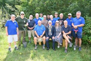 A group of fifteen people are gathered outdoors in a sunny, green area surrounded by trees and grass. Most of them are standing, while three people sit on a wooden bench at the front. The group consists of mostly older men and one woman. Several people are wearing bright blue polo shirts with a small green-and-yellow circular logo, suggesting they are part of a community or volunteer group.Some of the standing men are holding garden spades or shovels, and most are dressed casually in shorts, t-shirts, and outdoor shoes. The seated man in the middle wears a dark suit with a patterned tie and a ceremonial gold chain around his neck, indicating he may be a local mayor or official. The woman next to him wears a patterned dress and sunglasses, smiling toward the camera. Everyone appears cheerful and relaxed, enjoying the moment in what looks like a community or conservation project in a natural setting.