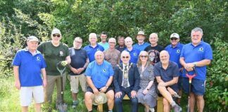 A group of fifteen people are gathered outdoors in a sunny, green area surrounded by trees and grass. Most of them are standing, while three people sit on a wooden bench at the front. The group consists of mostly older men and one woman. Several people are wearing bright blue polo shirts with a small green-and-yellow circular logo, suggesting they are part of a community or volunteer group. Some of the standing men are holding garden spades or shovels, and most are dressed casually in shorts, t-shirts, and outdoor shoes. The seated man in the middle wears a dark suit with a patterned tie and a ceremonial gold chain around his neck, indicating he may be a local mayor or official. The woman next to him wears a patterned dress and sunglasses, smiling toward the camera. Everyone appears cheerful and relaxed, enjoying the moment in what looks like a community or conservation project in a natural setting.