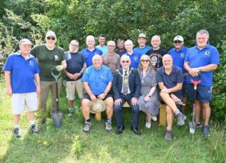 A group of fifteen people are gathered outdoors in a sunny, green area surrounded by trees and grass. Most of them are standing, while three people sit on a wooden bench at the front. The group consists of mostly older men and one woman. Several people are wearing bright blue polo shirts with a small green-and-yellow circular logo, suggesting they are part of a community or volunteer group. Some of the standing men are holding garden spades or shovels, and most are dressed casually in shorts, t-shirts, and outdoor shoes. The seated man in the middle wears a dark suit with a patterned tie and a ceremonial gold chain around his neck, indicating he may be a local mayor or official. The woman next to him wears a patterned dress and sunglasses, smiling toward the camera. Everyone appears cheerful and relaxed, enjoying the moment in what looks like a community or conservation project in a natural setting.