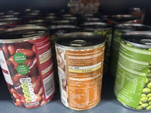 The image shows several metal cans of food arranged closely together on a supermarket shelf. The cans are different varieties of vegetables and beans. The lighting is typical of a grocery store aisle, and the viewpoint is close-up, focusing mainly on the labels and tops of the cans.