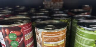 The image shows several metal cans of food arranged closely together on a supermarket shelf. The cans are different varieties of vegetables and beans. The lighting is typical of a grocery store aisle, and the viewpoint is close-up, focusing mainly on the labels and tops of the cans.
