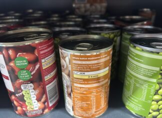 The image shows several metal cans of food arranged closely together on a supermarket shelf. The cans are different varieties of vegetables and beans. The lighting is typical of a grocery store aisle, and the viewpoint is close-up, focusing mainly on the labels and tops of the cans.