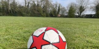 This photo shows a football sitting on short green grass in an open field. The ball is white with big red star shapes and looks a bit scuffed and used.