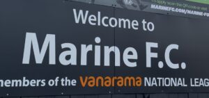 The image features a large, rectangular sign welcoming visitors to a Marine Football Club. It’s mounted prominently, near the entrance to the stadium. The sign reads: Welcome to Marine F.C. members of the Vanarama National League