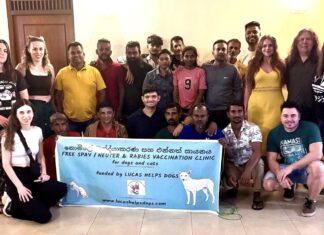 A group of 23 people stand and crouch around a banner advertising the Lucas Helps Dogs spay and neuter clinic.