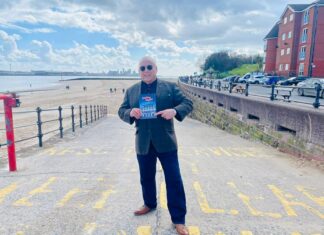 Paul Chase stands on the entrance to a beach holding a copy of his book 'Chelsea Nights'